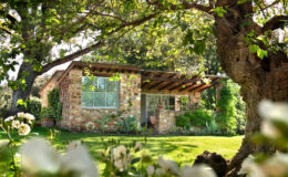 Charming stone cottage surrounded by lush greenery and flowers, featuring large windows and a wooden overhang, set in a serene garden environment.