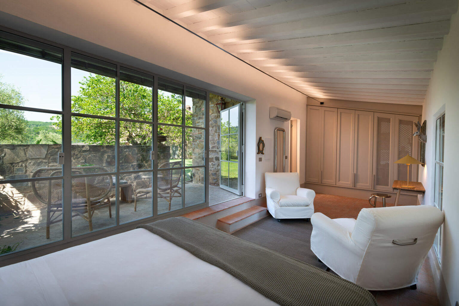 Bright and modern bedroom featuring a comfortable white armchair, a large bed with a textured blanket, and expansive glass doors leading to a scenic outdoor patio with stone walls and greenery. The room has a minimalist design with light wooden beams and stylish decor, creating a serene and inviting atmosphere. | Only on SopranoVillas