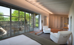 Bright and modern bedroom featuring a comfortable white armchair, a large bed with a textured blanket, and expansive glass doors leading to a scenic outdoor patio with stone walls and greenery. The room has a minimalist design with light wooden beams and stylish decor, creating a serene and inviting atmosphere.