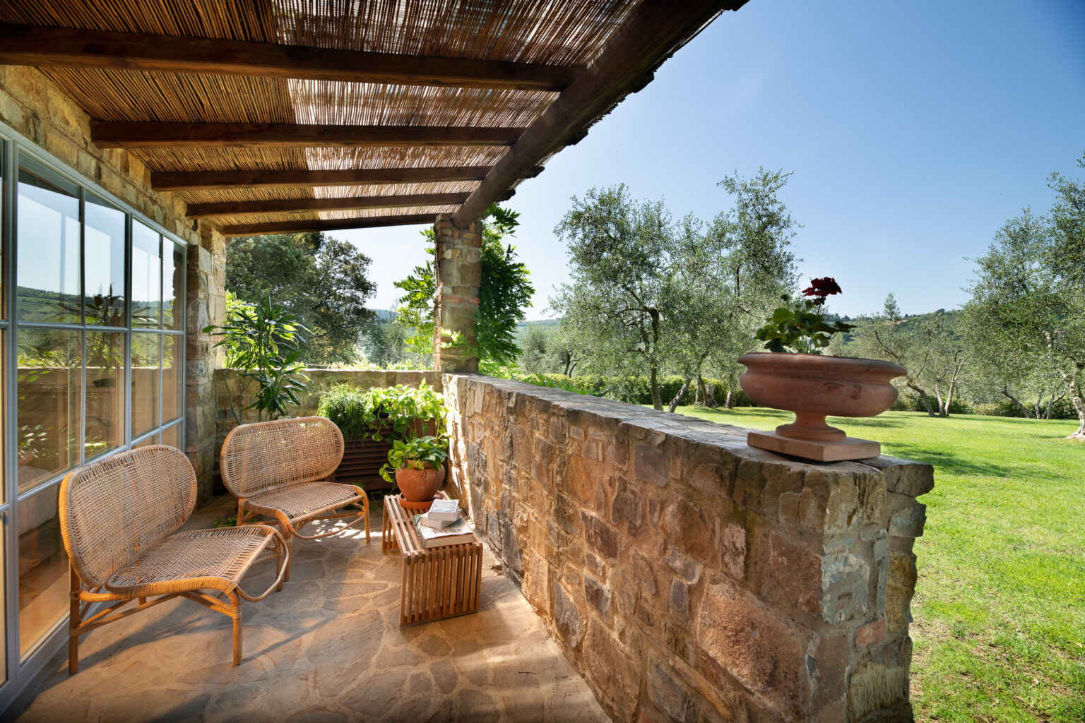 Cozy outdoor seating area with rattan chairs and a wooden table, surrounded by lush greenery and olive trees, under a shaded pergola. Perfect for relaxing in a serene garden setting. | Only on SopranoVillas