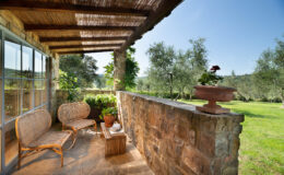 Cozy outdoor seating area with rattan chairs and a wooden table, surrounded by lush greenery and olive trees, under a shaded pergola. Perfect for relaxing in a serene garden setting.