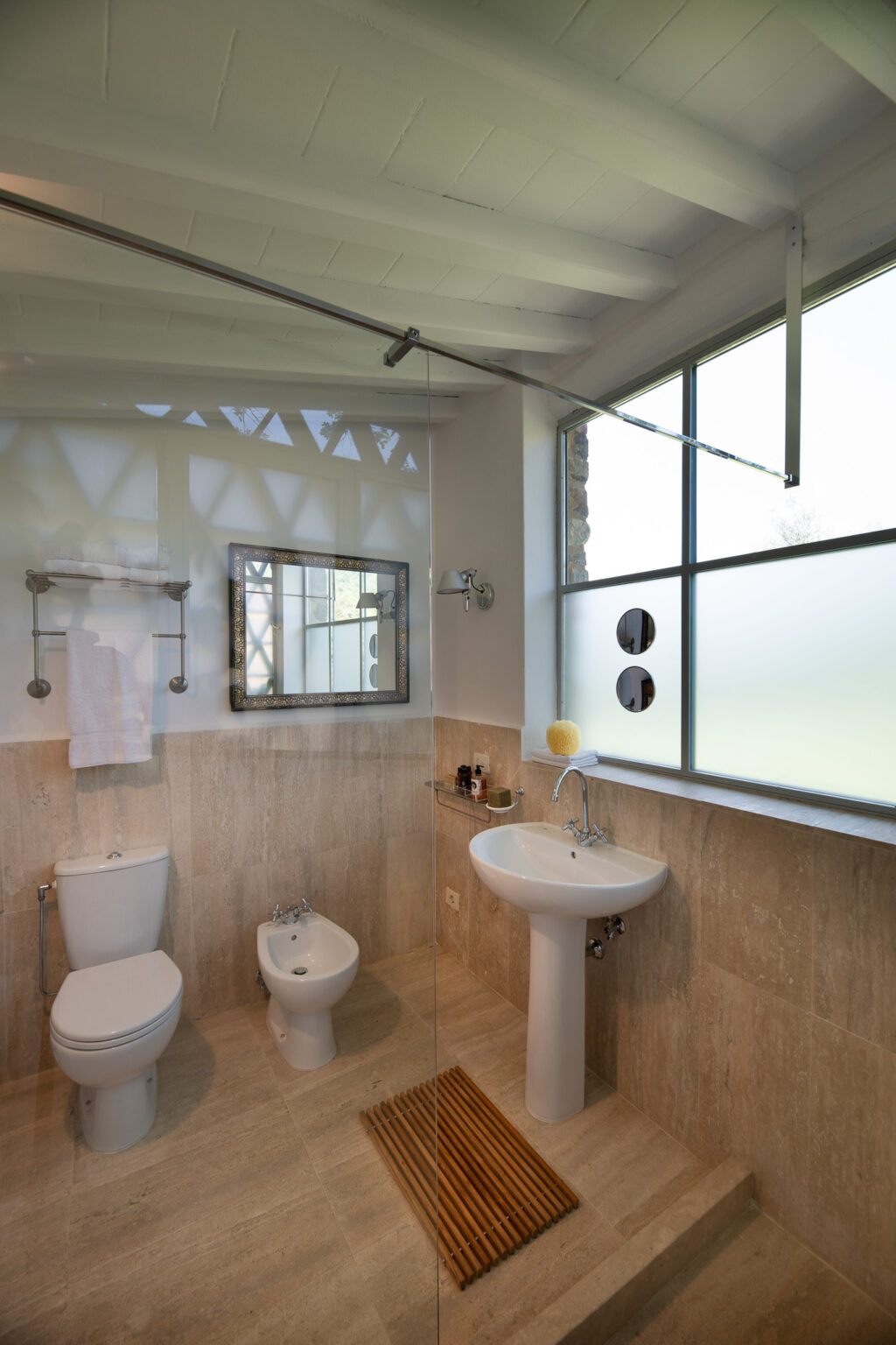 Modern bathroom featuring a glass shower, toilet, bidet, and a stylish sink with a wooden mat. Natural light streams in through large windows, highlighting the stone walls and elegant mirror. | Only on SopranoVillas