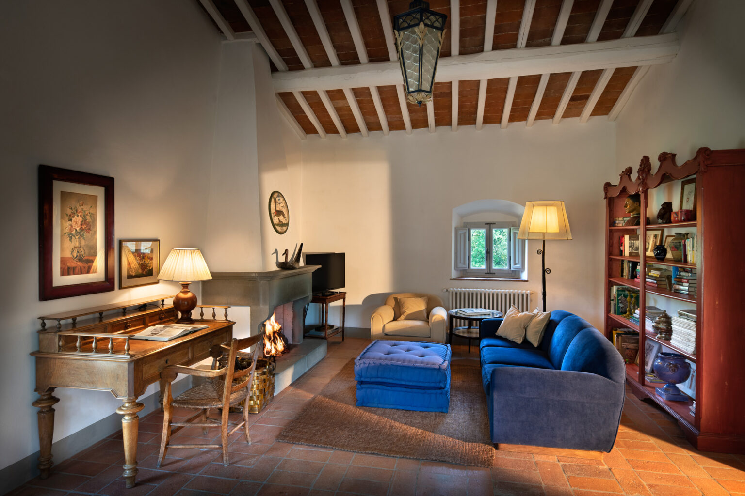 Cozy living room with wooden beams, featuring a blue sofa, an armchair, and a wooden desk. The room includes a modern TV, a decorative bookshelf, and a fireplace with a warm fire, creating an inviting atmosphere. Natural light streams in through a window, enhancing the rustic charm. | Only on SopranoVillas