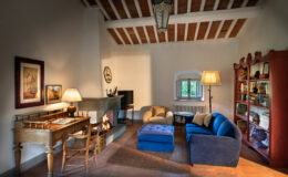 Cozy living room with wooden beams, featuring a blue sofa, an armchair, and a wooden desk. The room includes a modern TV, a decorative bookshelf, and a fireplace with a warm fire, creating an inviting atmosphere. Natural light streams in through a window, enhancing the rustic charm.