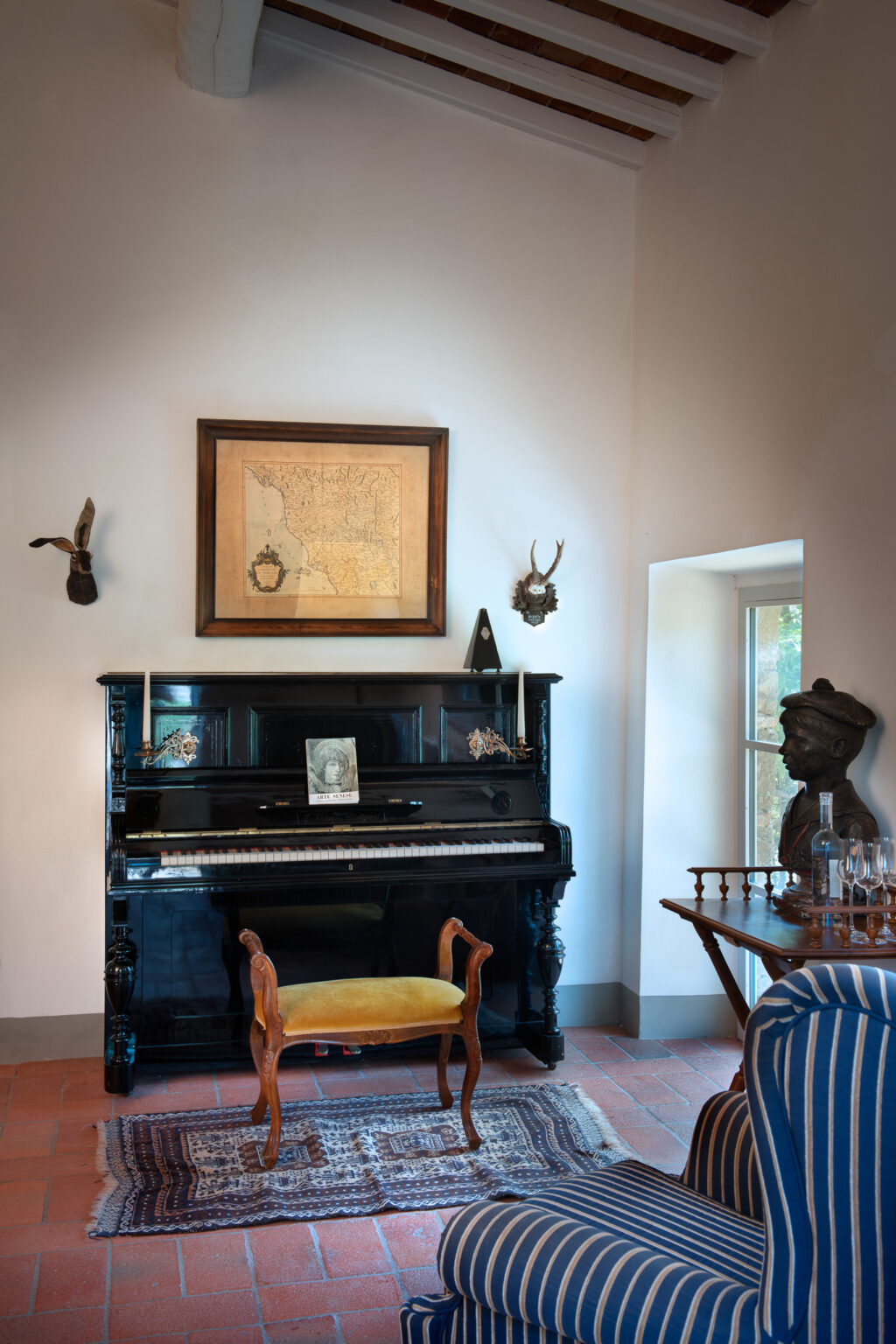 Elegant interior featuring a vintage black piano adorned with decorative elements, a framed map on the wall, and various antique wall mounts. A plush yellow stool is positioned in front of the piano on a patterned rug, while a wooden table with glassware and a bust sculpture adds charm to the cozy space. | Only on SopranoVillas