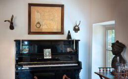 Elegant interior featuring a vintage black piano adorned with decorative elements, a framed map on the wall, and various antique wall mounts. A plush yellow stool is positioned in front of the piano on a patterned rug, while a wooden table with glassware and a bust sculpture adds charm to the cozy space.