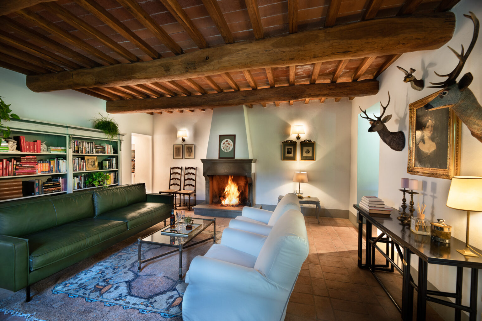 Cozy living room featuring a green leather sofa and white armchairs, a rustic wooden beam ceiling, and a stone fireplace with a warm fire. Decor includes a bookshelf filled with colorful books, framed art on the walls, and decorative taxidermy. A glass coffee table sits on a patterned rug, creating an inviting atmosphere. | Only on SopranoVillas