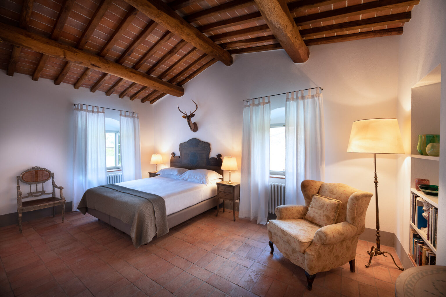 Cozy bedroom featuring a queen-sized bed with a neutral-toned blanket, wooden beams on the ceiling, and large windows draped with sheer curtains. The room includes a vintage armchair, bedside lamps, and a decorative wall mount, creating a warm and inviting atmosphere. Ideal for relaxation and restful stays. | Only on SopranoVillas