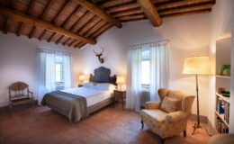 Cozy bedroom featuring a queen-sized bed with a neutral-toned blanket, wooden beams on the ceiling, and large windows draped with sheer curtains. The room includes a vintage armchair, bedside lamps, and a decorative wall mount, creating a warm and inviting atmosphere. Ideal for relaxation and restful stays.