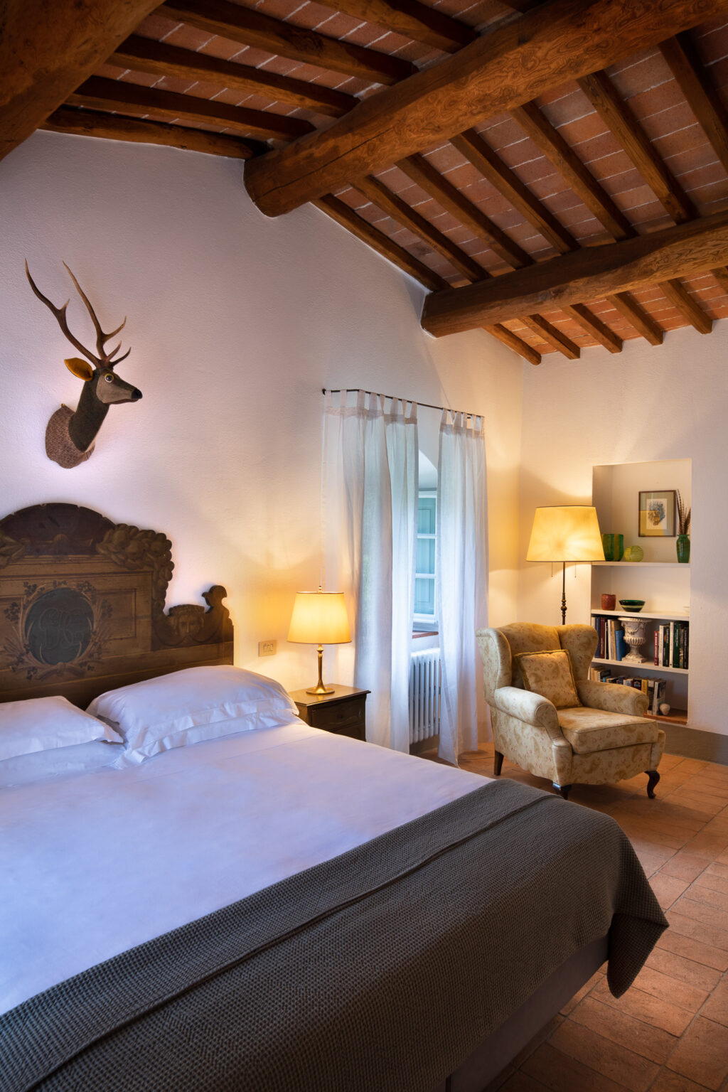 Cozy bedroom featuring a wooden bed with white linens, a decorative deer head on the wall, and a comfortable armchair. The room has a rustic wooden ceiling, soft lighting from a bedside lamp, and sheer curtains allowing natural light to enter. A small bookshelf adds a touch of charm to the inviting space. | Only on SopranoVillas