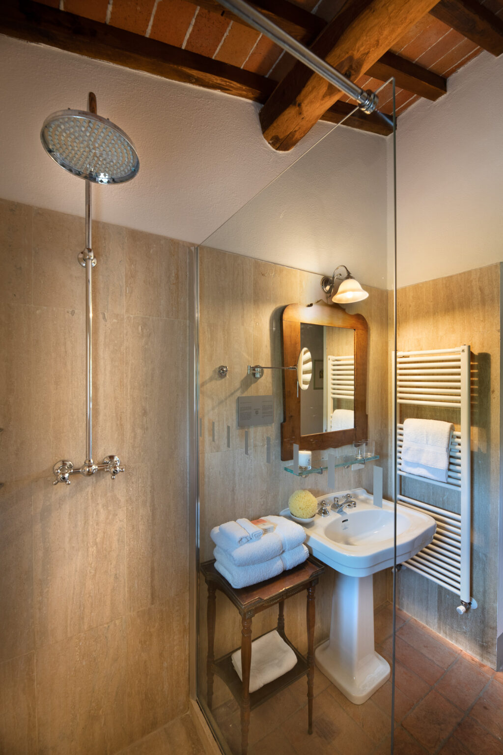 Modern bathroom featuring a spacious shower with a rain showerhead, a stylish sink with a vintage mirror, and neatly arranged towels on a wooden stool. The warm-toned tiles and wooden beams create a cozy atmosphere. | Only on SopranoVillas