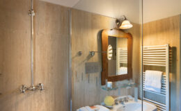 Modern bathroom featuring a spacious shower with a rain showerhead, a stylish sink with a vintage mirror, and neatly arranged towels on a wooden stool. The warm-toned tiles and wooden beams create a cozy atmosphere.