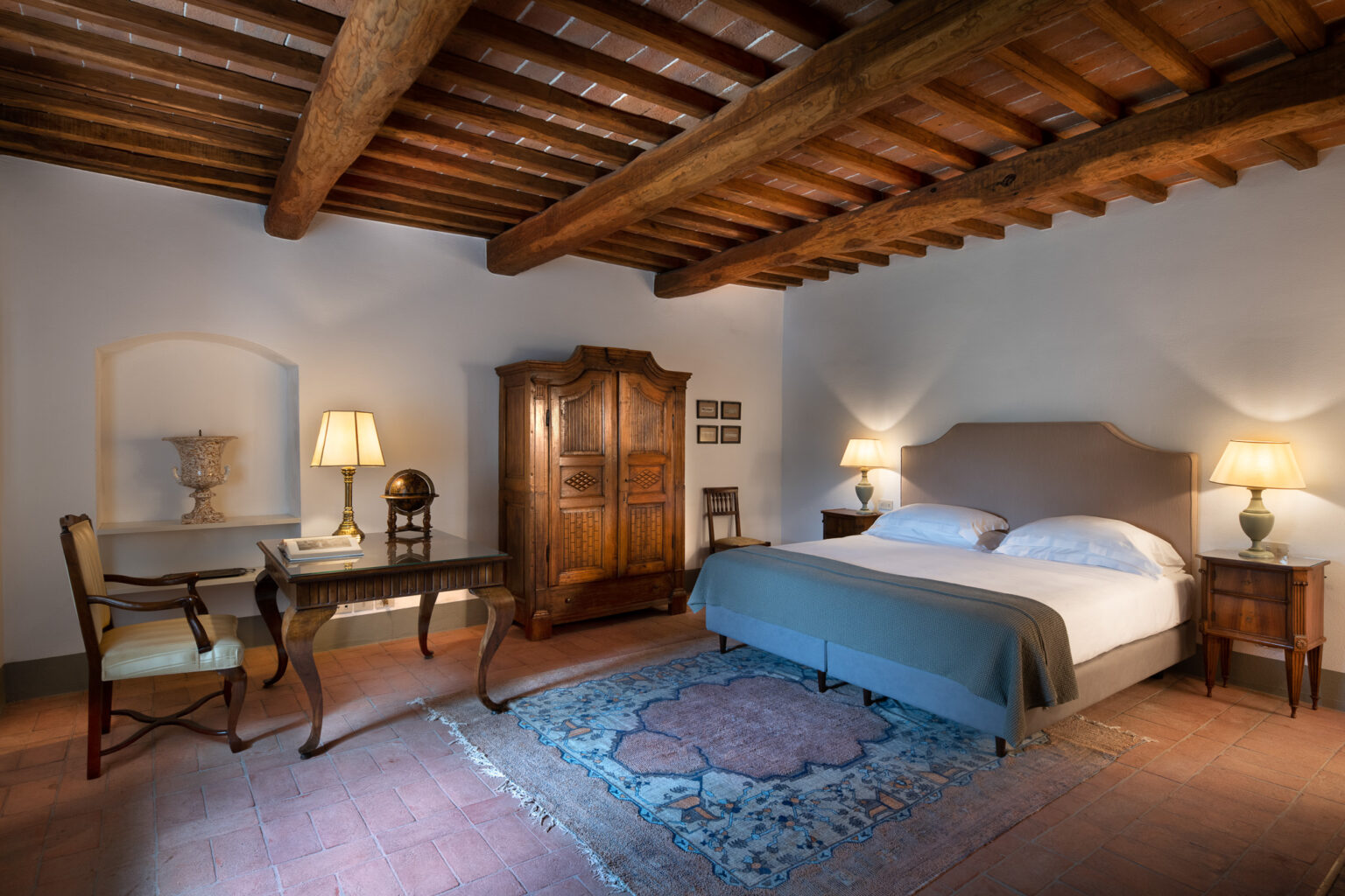 Cozy bedroom featuring rustic wooden beams, a comfortable double bed with soft linens, an elegant desk with a chair, and antique furnishings including a wardrobe and decorative globe. Warm lighting creates a serene atmosphere, complemented by a traditional area rug. Ideal for a relaxing getaway. | Only on SopranoVillas