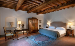 Cozy bedroom featuring rustic wooden beams, a comfortable double bed with soft linens, an elegant desk with a chair, and antique furnishings including a wardrobe and decorative globe. Warm lighting creates a serene atmosphere, complemented by a traditional area rug. Ideal for a relaxing getaway.