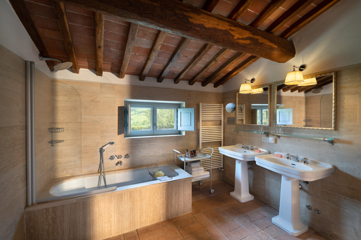 Spacious and elegantly designed bathroom featuring a bathtub, two sinks, warm wooden beams, and a picturesque window with a view of the countryside. The room includes modern fixtures, a towel rack, and a stylish mirror, creating a relaxing atmosphere. | Only on SopranoVillas