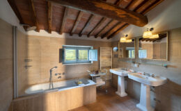 Spacious and elegantly designed bathroom featuring a bathtub, two sinks, warm wooden beams, and a picturesque window with a view of the countryside. The room includes modern fixtures, a towel rack, and a stylish mirror, creating a relaxing atmosphere.