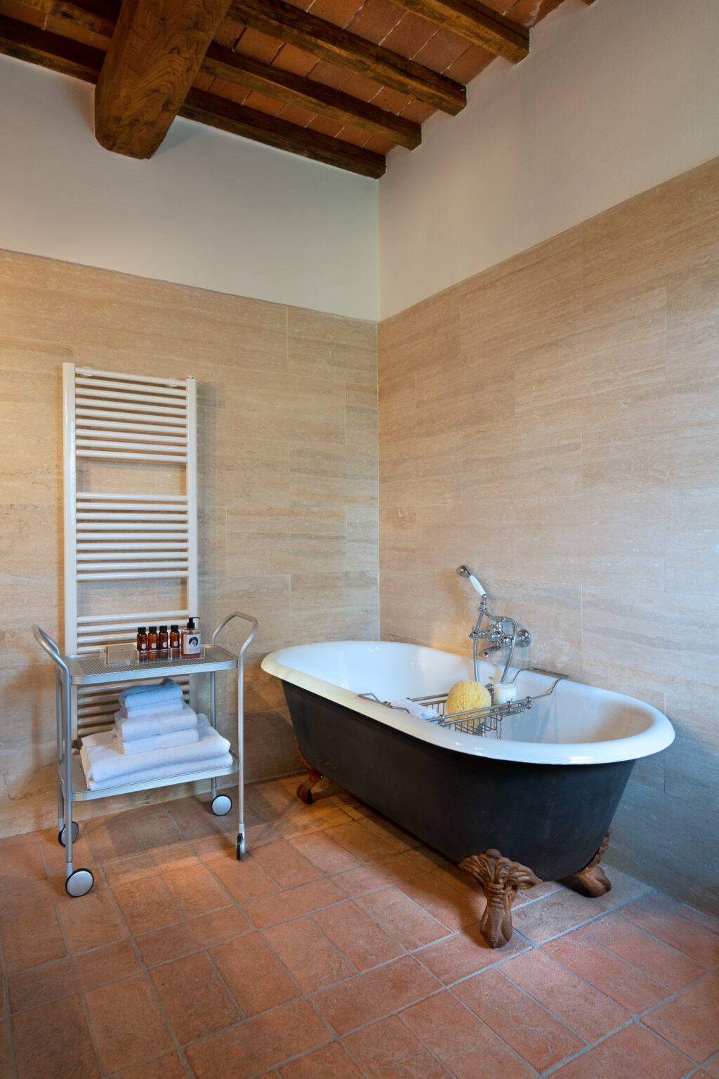 Elegant bathroom featuring a vintage clawfoot bathtub, a modern towel rack, and neatly stacked white towels on a metal cart. The warm terracotta floor and natural stone walls create a cozy atmosphere, complemented by wooden beams on the ceiling. Ideal for relaxation and spa-like experiences. | Only on SopranoVillas