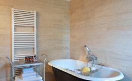Elegant bathroom featuring a vintage clawfoot bathtub, a modern towel rack, and neatly stacked white towels on a metal cart. The warm terracotta floor and natural stone walls create a cozy atmosphere, complemented by wooden beams on the ceiling. Ideal for relaxation and spa-like experiences.