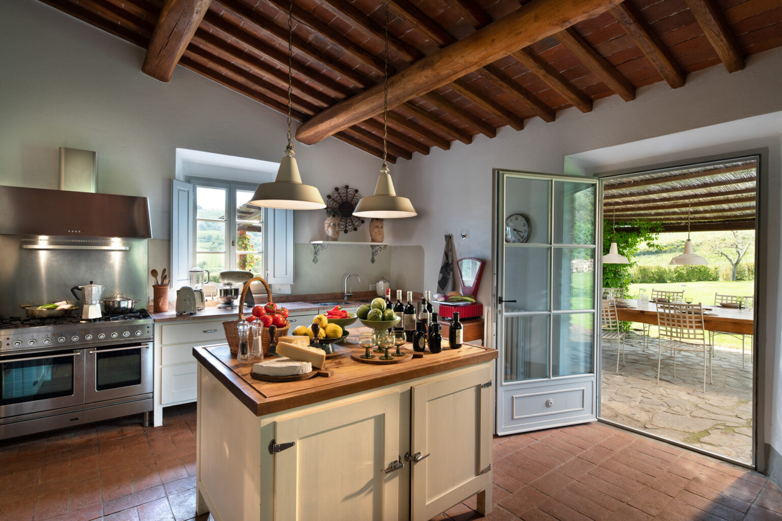 Bright and spacious rustic kitchen featuring a wooden ceiling, modern appliances, and a central island adorned with fresh fruits and cheese. The open door leads to a scenic outdoor dining area, creating a seamless connection between indoor and outdoor living spaces. Ideal for culinary enthusiasts and entertaining. | Only on SopranoVillas