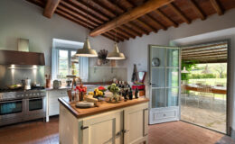 Bright and spacious rustic kitchen featuring a wooden ceiling, modern appliances, and a central island adorned with fresh fruits and cheese. The open door leads to a scenic outdoor dining area, creating a seamless connection between indoor and outdoor living spaces. Ideal for culinary enthusiasts and entertaining.