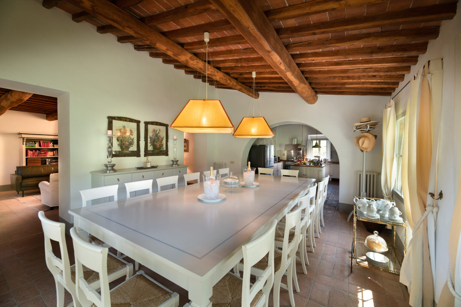 Spacious dining area featuring a long white table surrounded by wooden chairs, illuminated by stylish pendant lights. The rustic charm is enhanced by exposed wooden beams and decorative paintings on the walls. A glimpse of a modern kitchen and cozy living space can be seen in the background. | Only on SopranoVillas