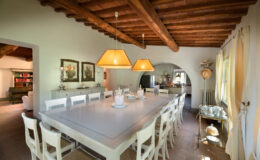 Spacious dining area featuring a long white table surrounded by wooden chairs, illuminated by stylish pendant lights. The rustic charm is enhanced by exposed wooden beams and decorative paintings on the walls. A glimpse of a modern kitchen and cozy living space can be seen in the background.
