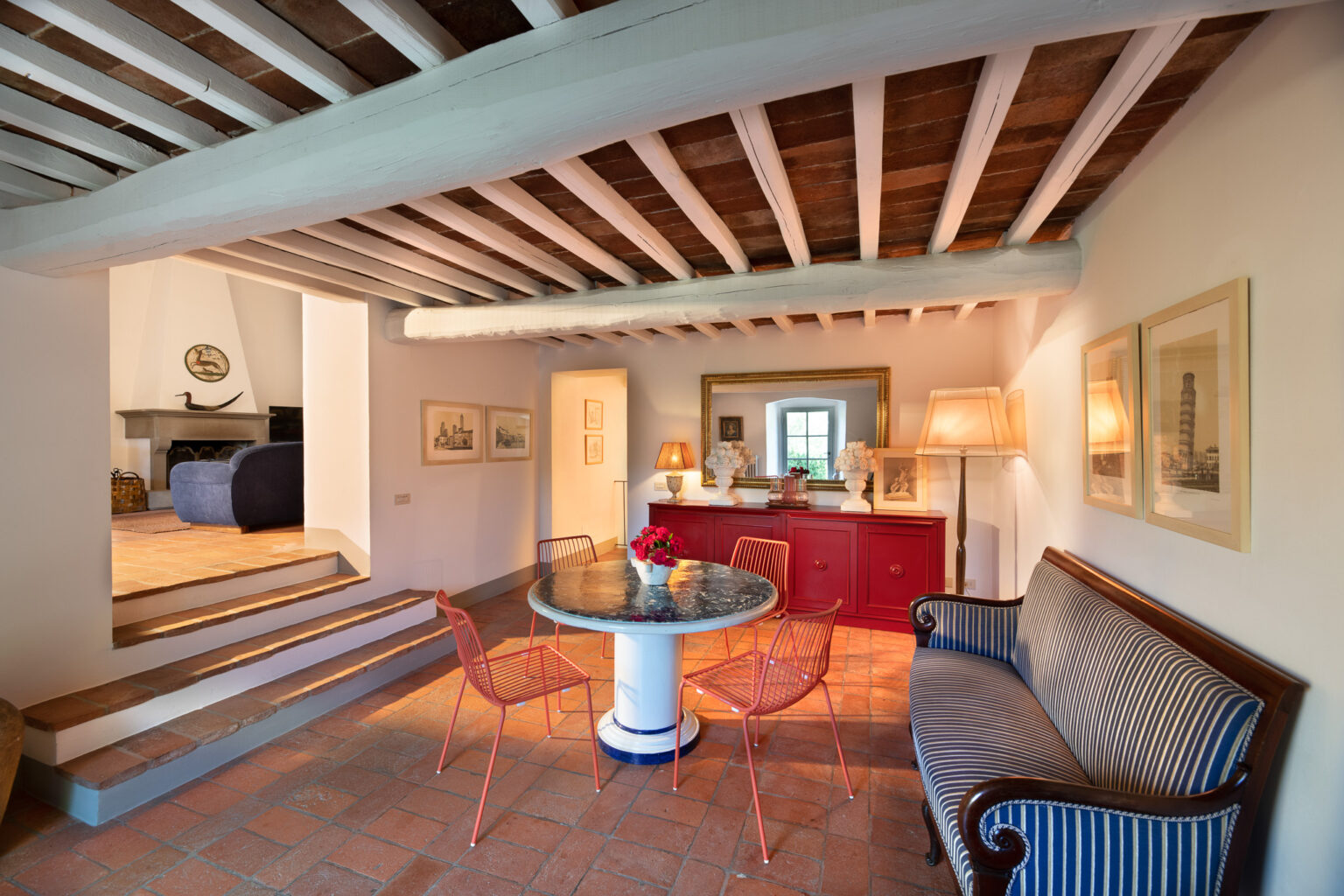 Cozy interior of a stylish living space featuring a round marble dining table surrounded by red wire chairs, a blue striped sofa, and a red sideboard. The room showcases exposed wooden beams and terracotta flooring, with decorative artwork and lamps accentuating the warm ambiance. | Only on SopranoVillas