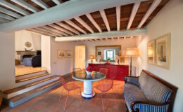 Cozy interior of a stylish living space featuring a round marble dining table surrounded by red wire chairs, a blue striped sofa, and a red sideboard. The room showcases exposed wooden beams and terracotta flooring, with decorative artwork and lamps accentuating the warm ambiance.