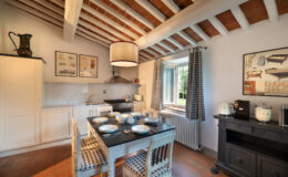 Modern kitchen interior with wooden beam ceiling, featuring a dining table set for six, stylish appliances, and decorative artwork on the walls. Natural light streams in through a window, enhancing the inviting atmosphere.