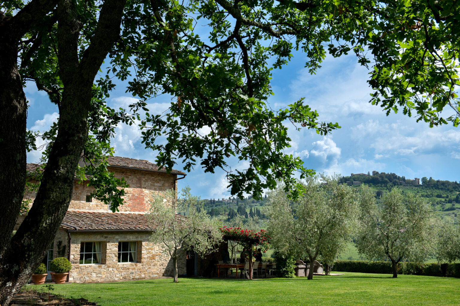 A picturesque countryside scene featuring a rustic stone house surrounded by lush greenery, olive trees, and a vibrant garden. The background showcases rolling hills and a blue sky with fluffy clouds, creating a serene and inviting atmosphere. Ideal for promoting rural tourism or vacation rentals in scenic locations. | Only on SopranoVillas