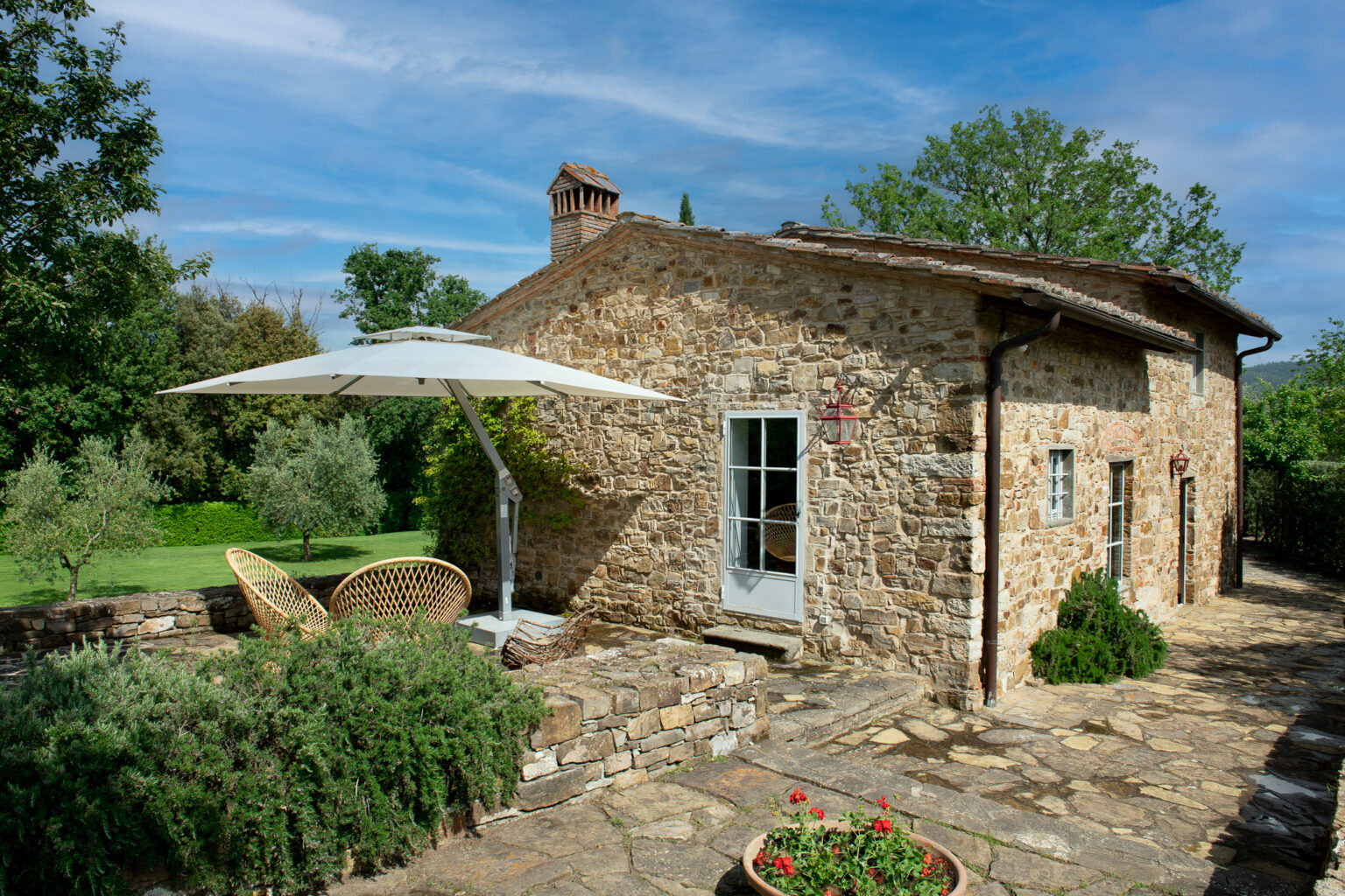 Charming stone cottage with a patio, featuring a large umbrella and wicker chairs, surrounded by lush greenery and flowering plants, under a blue sky. Ideal for outdoor relaxation and enjoying nature. | Only on SopranoVillas
