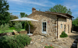 Charming stone cottage with a patio, featuring a large umbrella and wicker chairs, surrounded by lush greenery and flowering plants, under a blue sky. Ideal for outdoor relaxation and enjoying nature.