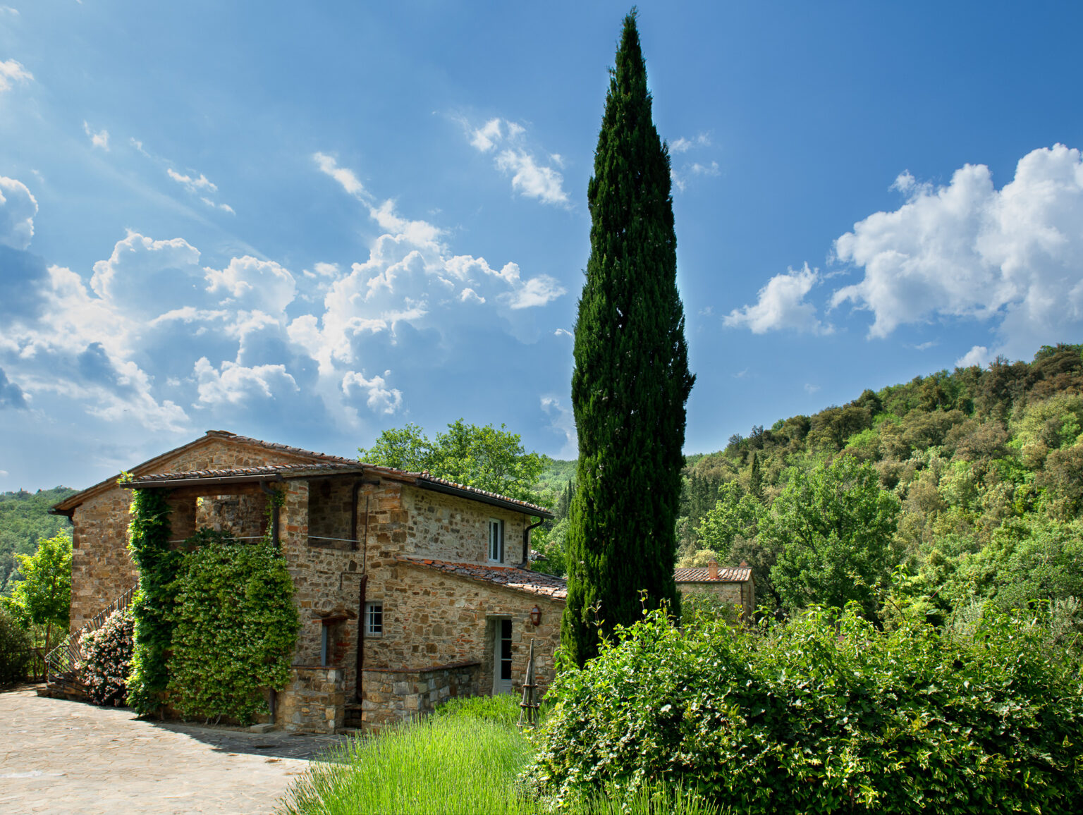 Rustic stone house surrounded by lush greenery and a tall cypress tree, under a bright blue sky with fluffy clouds. Ideal for nature lovers and those seeking a tranquil getaway in the countryside. | Only on SopranoVillas