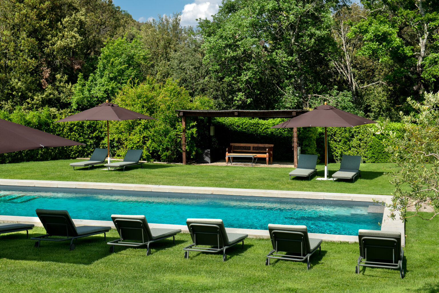 Luxurious outdoor pool area featuring comfortable lounge chairs, shaded by large umbrellas, surrounded by lush greenery and a cozy seating area under a pergola. Ideal for relaxation and leisure. | Only on SopranoVillas