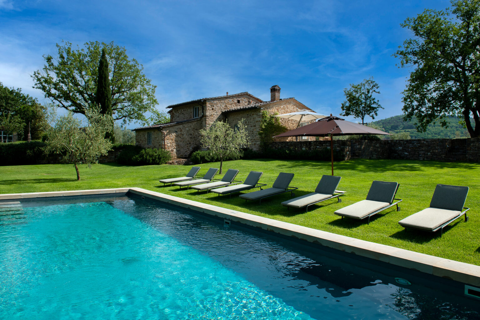 Serene outdoor setting featuring a sparkling swimming pool, lined with modern lounge chairs on lush green grass, with a rustic stone house and trees in the background under a clear blue sky. Perfect for relaxation and leisure. | Only on SopranoVillas