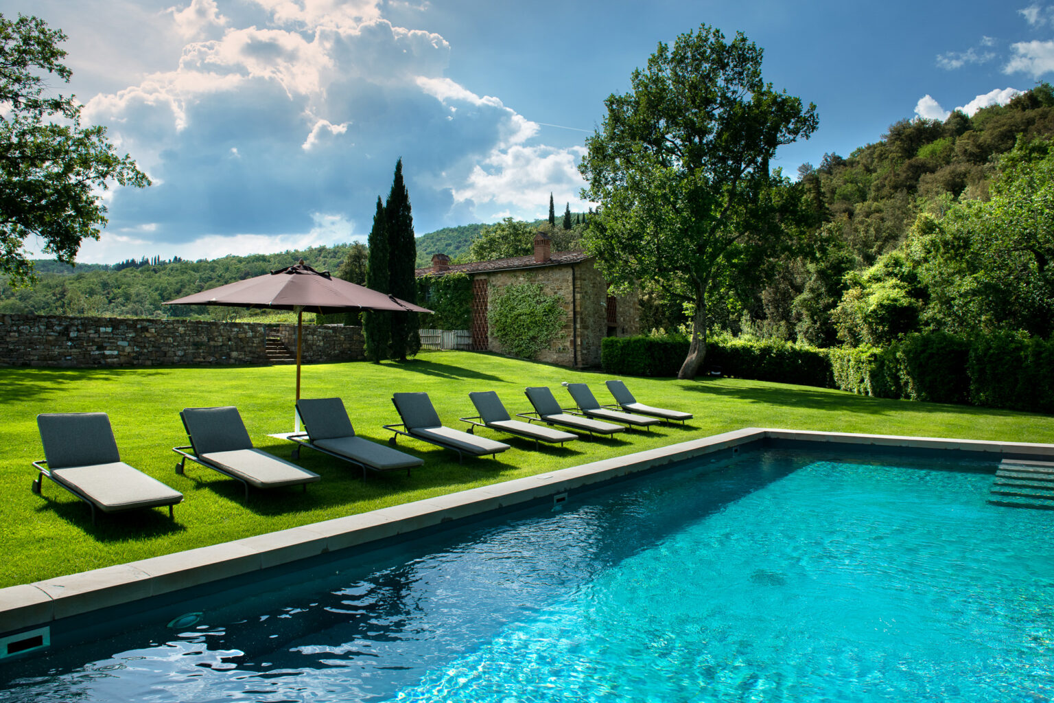 Luxury outdoor pool area featuring comfortable lounge chairs and a shaded umbrella, surrounded by lush greenery and scenic hills, perfect for relaxation and leisure. | Only on SopranoVillas