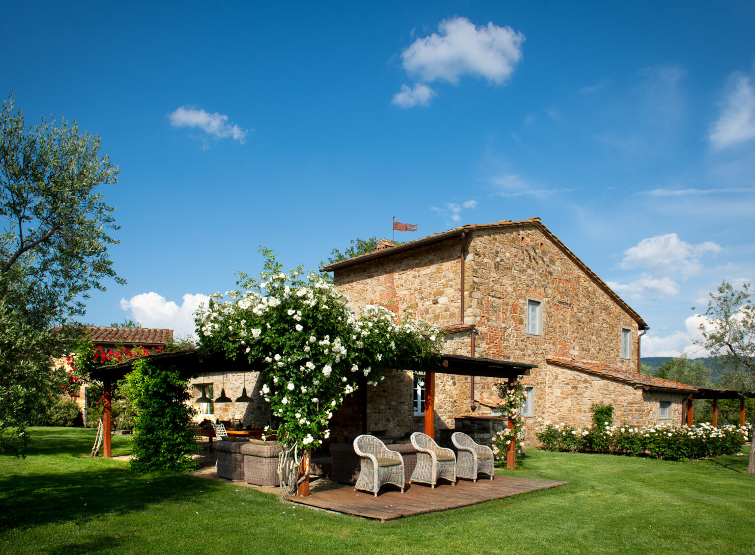 Charming stone villa surrounded by lush greenery and blooming white roses, featuring a wooden deck with comfortable wicker seating, under a clear blue sky. Ideal for outdoor relaxation and scenic countryside views. | Only on SopranoVillas
