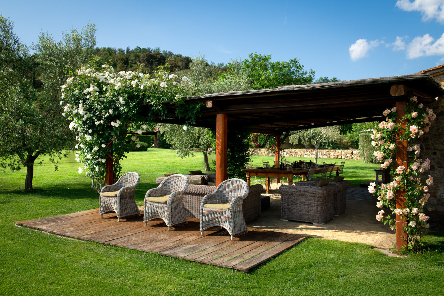 Cozy outdoor lounge area with wicker chairs and a wooden deck, surrounded by lush greenery and blooming white roses, under a pergola. Perfect for relaxation and gatherings in a serene garden setting. | Only on SopranoVillas