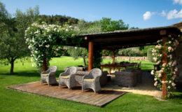 Cozy outdoor lounge area with wicker chairs and a wooden deck, surrounded by lush greenery and blooming white roses, under a pergola. Perfect for relaxation and gatherings in a serene garden setting.