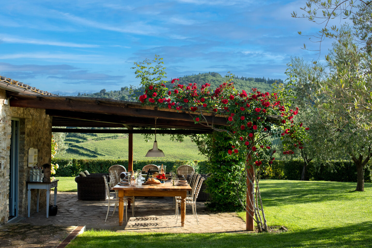 A scenic outdoor dining area surrounded by lush greenery and blooming red roses, featuring a wooden table set for a meal, wicker chairs, and a cozy ambiance with rolling hills in the background. Perfect for al fresco dining and relaxation in a tranquil countryside setting. | Only on SopranoVillas
