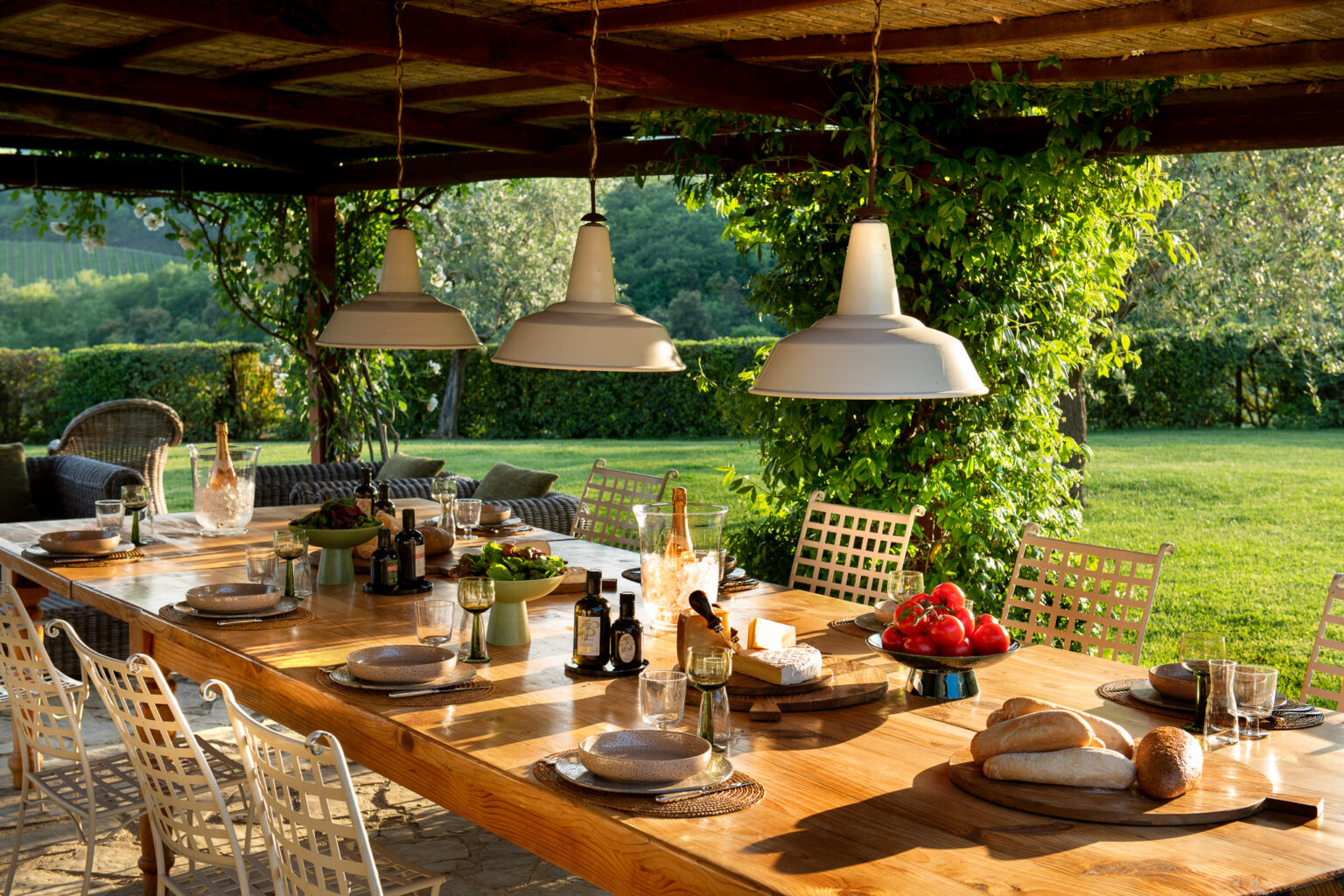 Outdoor dining area featuring a rustic wooden table set for a meal, adorned with plates, glasses, olive oil, and fresh ingredients like tomatoes and bread. The space is illuminated by stylish pendant lights, surrounded by lush greenery and comfortable seating, creating an inviting atmosphere for al fresco dining. | Only on SopranoVillas