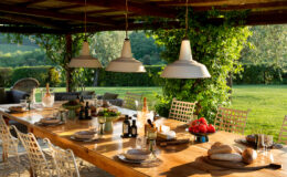 Outdoor dining area featuring a rustic wooden table set for a meal, adorned with plates, glasses, olive oil, and fresh ingredients like tomatoes and bread. The space is illuminated by stylish pendant lights, surrounded by lush greenery and comfortable seating, creating an inviting atmosphere for al fresco dining.