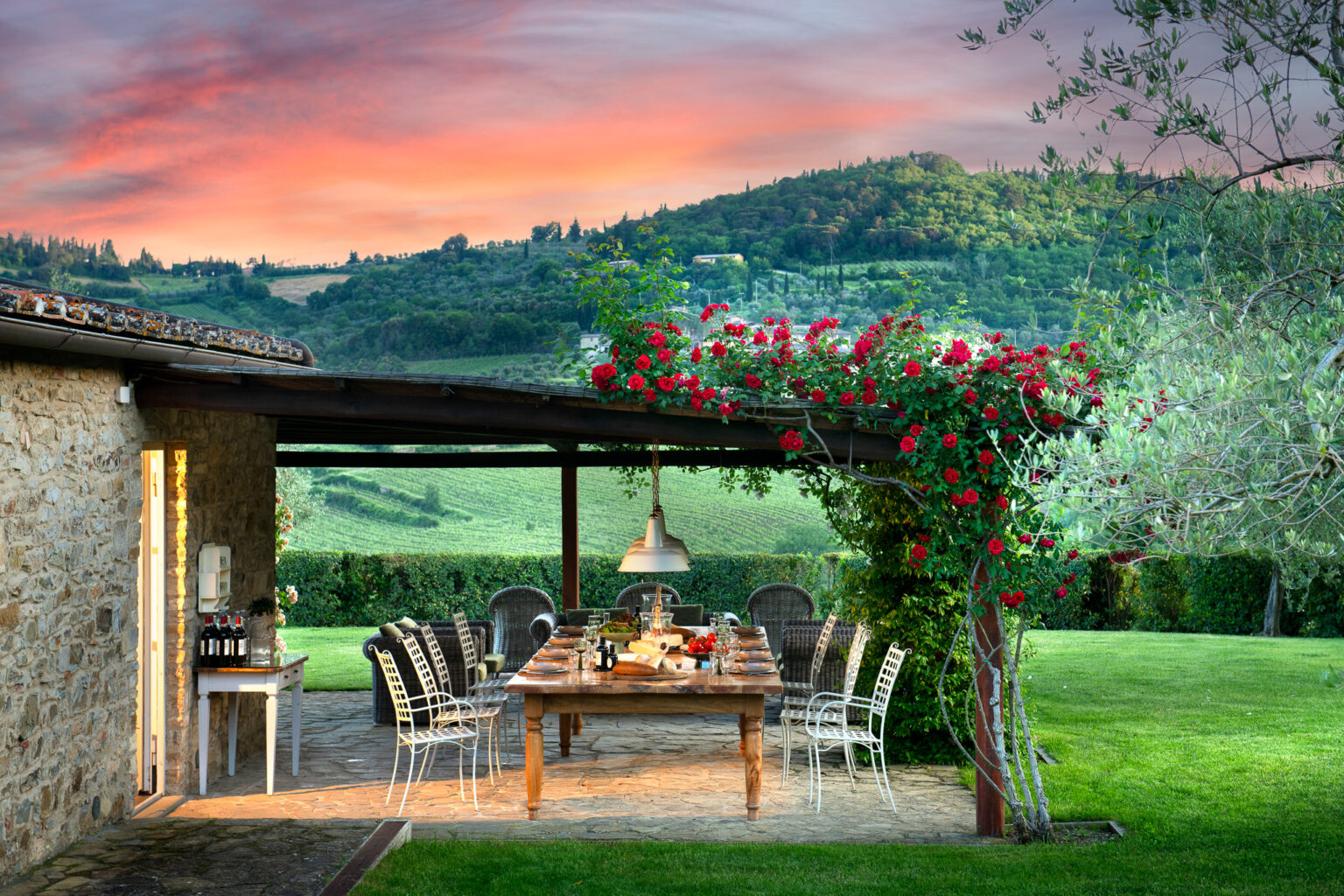 Outdoor dining area with a wooden table set for a meal, surrounded by elegant chairs, vibrant red roses, and lush green hills in the background during sunset. Ideal for gatherings and enjoying nature. | Only on SopranoVillas