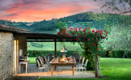 Outdoor dining area with a wooden table set for a meal, surrounded by elegant chairs, vibrant red roses, and lush green hills in the background during sunset. Ideal for gatherings and enjoying nature.