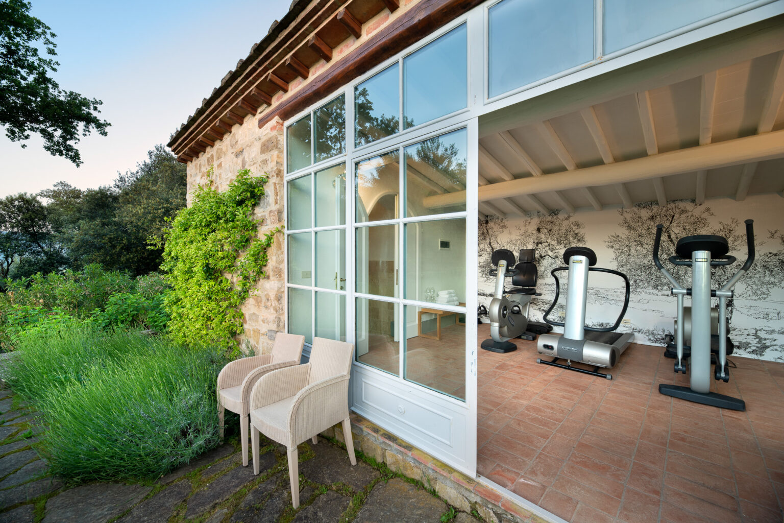 Modern home gym with exercise equipment visible through large glass windows, surrounded by lush greenery and stone pathways, featuring two outdoor chairs for relaxation. | Only on SopranoVillas