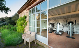 Modern home gym with exercise equipment visible through large glass windows, surrounded by lush greenery and stone pathways, featuring two outdoor chairs for relaxation.