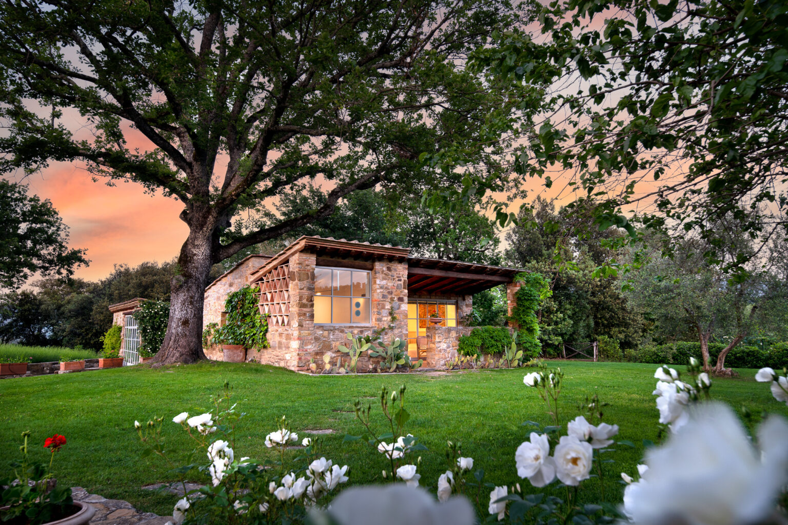 Charming stone cottage surrounded by lush greenery and blooming white flowers, set against a colorful sunset sky. The home features large windows and a rustic design, ideal for nature lovers seeking tranquility. | Only on SopranoVillas