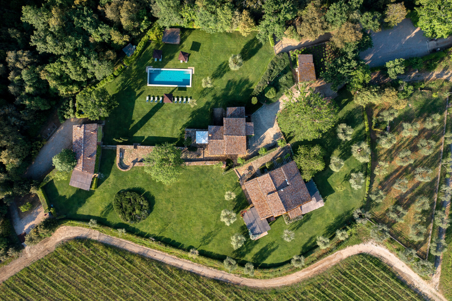 Aerial view of a luxurious countryside villa featuring a large green lawn, a swimming pool surrounded by lounge chairs, and multiple rustic buildings amidst lush trees and gardens. The property is bordered by a vineyard, showcasing the serene landscape of the area. | Only on SopranoVillas