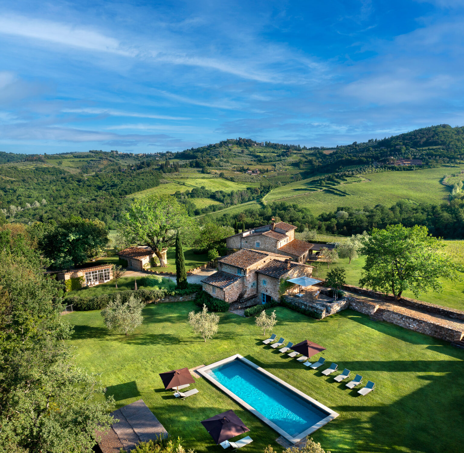 Aerial view of a rustic villa in Tuscany featuring a swimming pool, sun loungers, and lush green landscapes with rolling hills and vineyards in the background. Ideal for a relaxing getaway in the Italian countryside. | Only on SopranoVillas