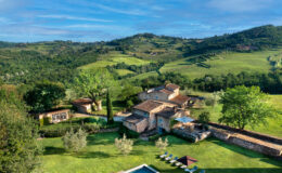 Aerial view of a rustic villa in Tuscany featuring a swimming pool, sun loungers, and lush green landscapes with rolling hills and vineyards in the background. Ideal for a relaxing getaway in the Italian countryside.